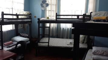 A dormitory room with bunk beds, a desk, a chair, and a fan.