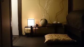 A dimly lit room with a bed, a bedside table with a lamp, a vase with branches, and a window with wooden shutters.