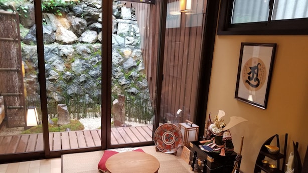 A room with tatami flooring, a wooden table, a cushioned bench, a small shelf with decorative items, and a view of a rocky garden through large glass windows. - Kyonoyado Senkakubettei (Kyoto)