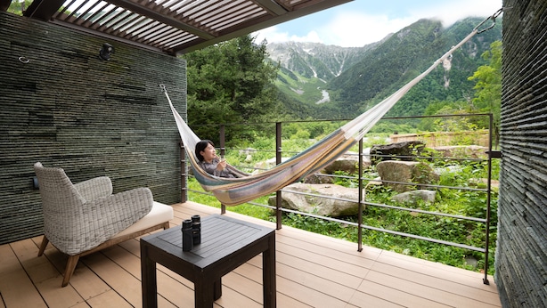 A hammock on a balcony with a mountain view, a cushioned chair, and a table with a bottle.