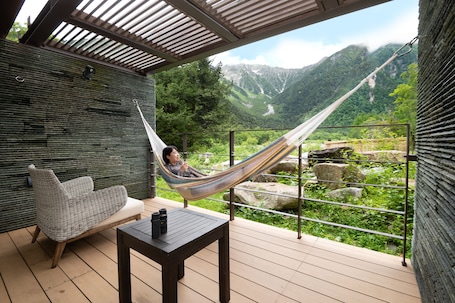 A hammock on a balcony with a mountain view, a cushioned chair, and a table with a bottle.