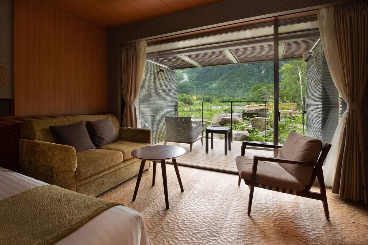 A room with a sofa, armchair, and small table, a large window with curtains, and a view of a mountainous landscape.