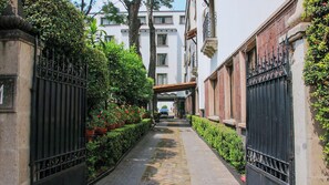 A narrow alleyway with a paved path, flanked by buildings with iron gates and lush greenery.