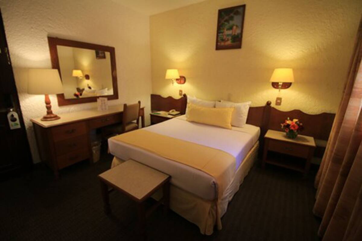A hotel room with a bed, bedside tables, a desk, a chair, a mirror, and lamps.