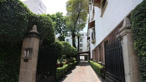 A narrow cobblestone pathway flanked by hedges and black metal gates, leading to a building with a white facade and a stone pillar marked with the number 31.