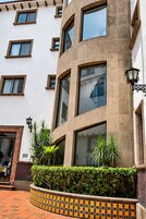 Un edificio de varios pisos con ventanas amplias, entrada con mosaicos y un pequeño jardín con plantas.