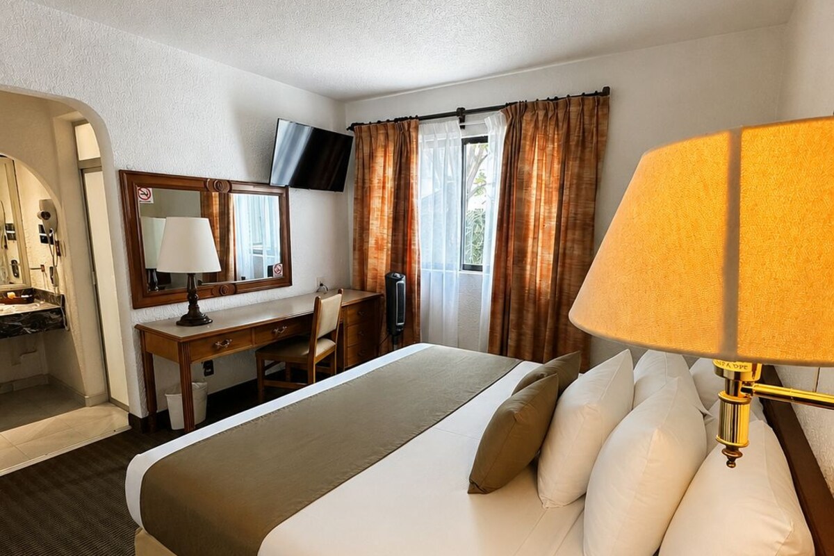 Habitación de hotel con cama, escritorio, silla, televisión y ventana con cortinas.