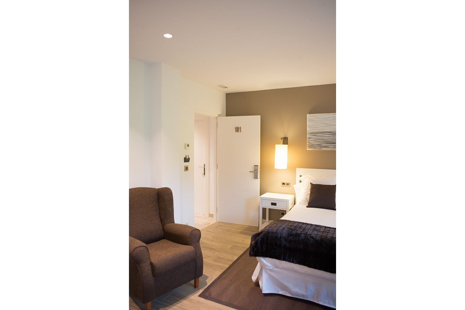 Une chambre d’hôtel avec un lit, une chaise, une table de chevet et une lampe fixée au mur.