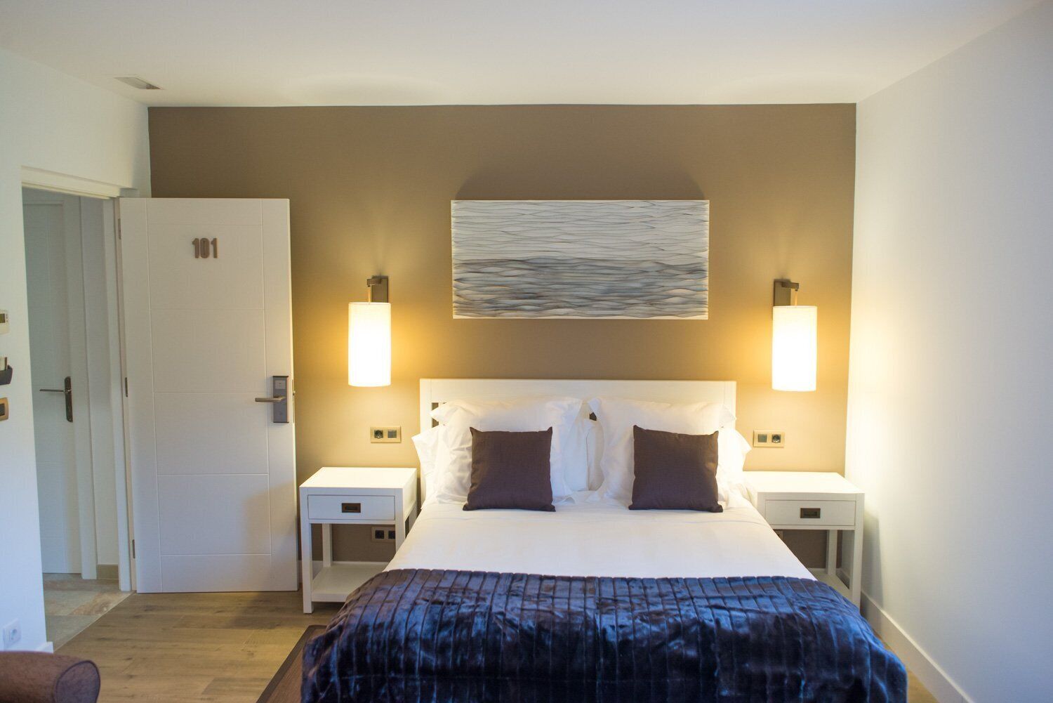 A hotel room with a large bed, two bedside tables, a wall-mounted artwork, and two wall sconces.