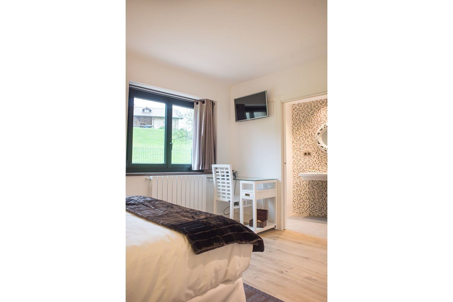 Un dormitorio con cama, escritorio, silla, televisor y baño con pared de azulejos y espejo.