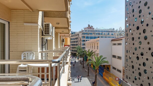 A balcony with a view of a city street, palm trees, and a building with circular windows. - Apartamentos Maja Beach (Benidorm)