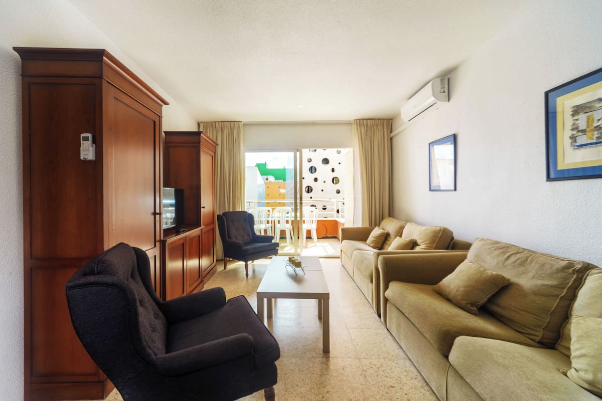 A living room with a sofa, armchairs, a coffee table, and a TV. There is a door with a lock and a window with curtains.