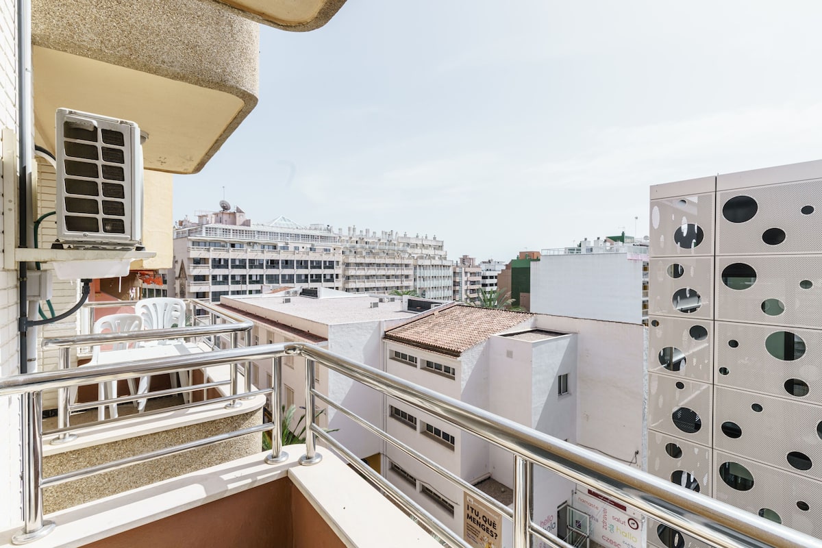 A balcony with a metal railing, a white air conditioning unit, and a view of a multi-story building with a tiled roof.
