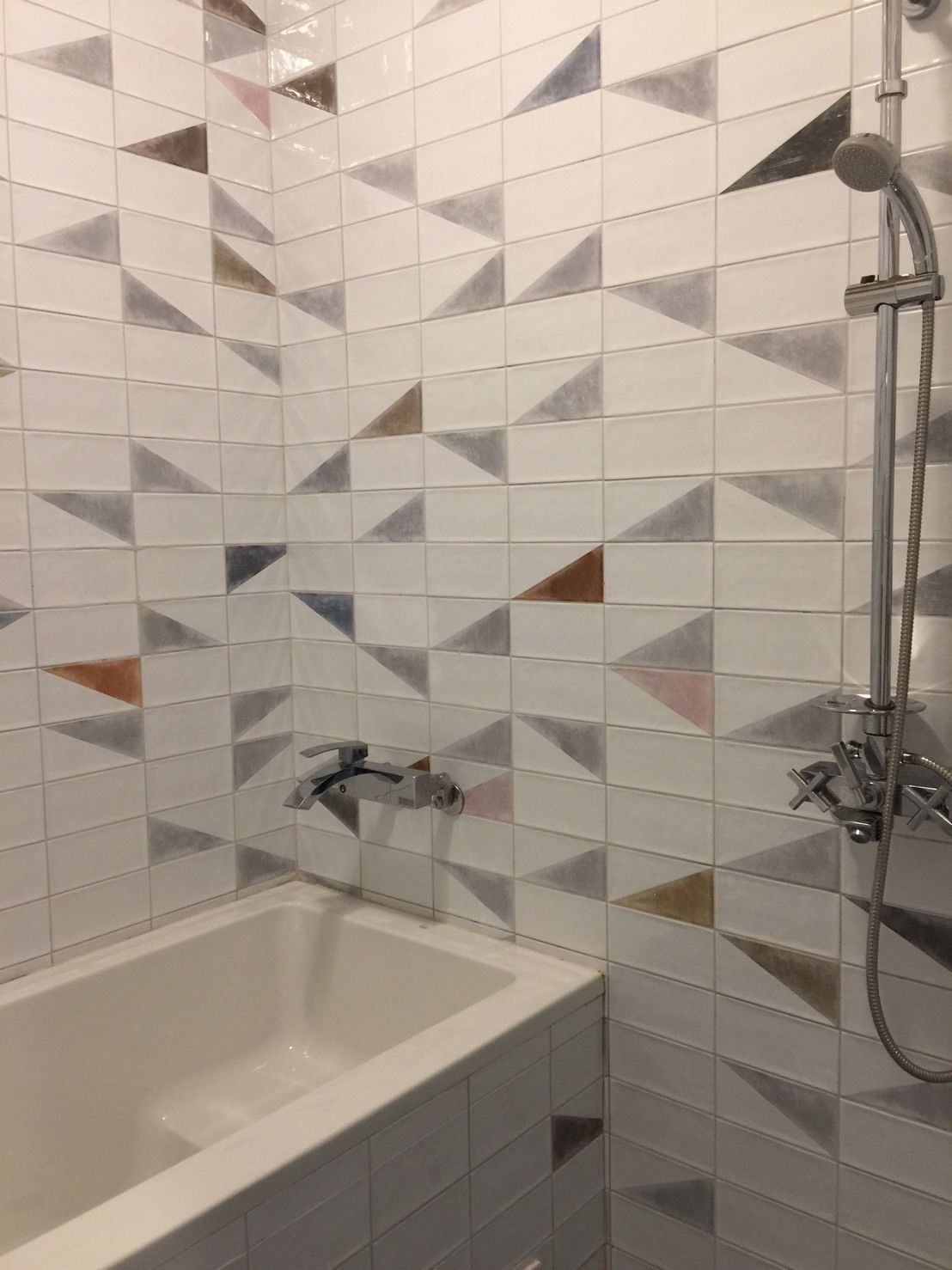 Une salle de bain avec une baignoire blanche, une douche équipée d’un pommeau de douche à main et un mur recouvert de carreaux aux motifs géométriques.