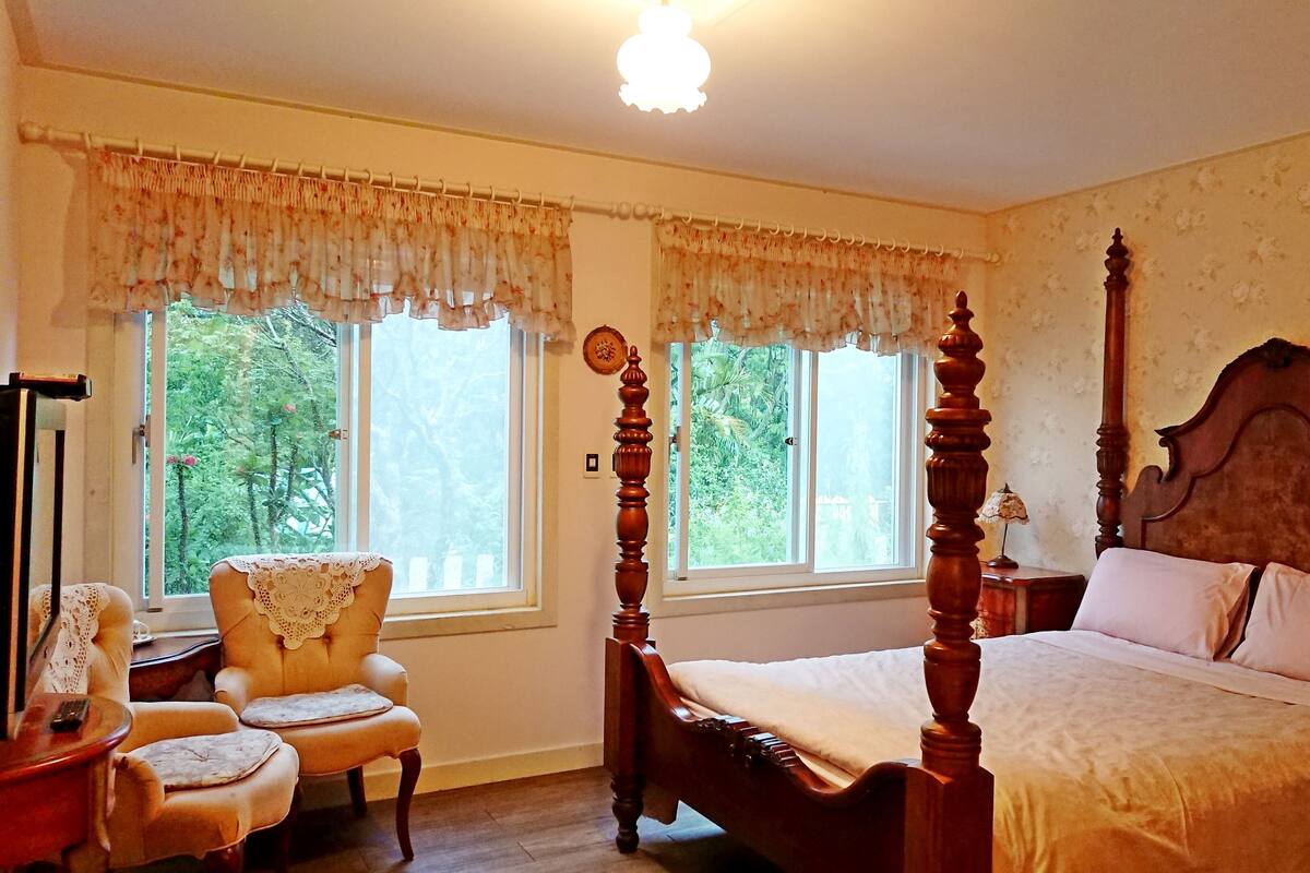Ein traditionell eingerichtetes Schlafzimmer mit Himmelbett, zwei Fenstern mit Vorhängen, einem Sessel und einem Nachttisch.