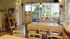 Un coin repas avec une table en verre et des chaises blanches, une étagère en forme d’échelle ornée de plantes, et une vue sur la verdure à l’extérieur.