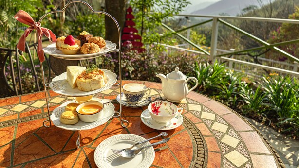 Un présentoir à trois niveaux, garni de pâtisseries, et une tasse de thé, le tout posé sur une table carrelée à l’extérieur, offrant une vue panoramique.