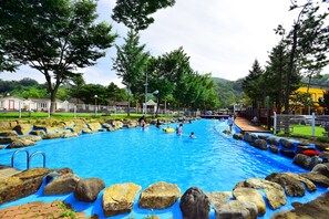 Ein Schwimmbecken mit Steinen und einer Holzterrasse, umgeben von Bäumen und einem Zaun.