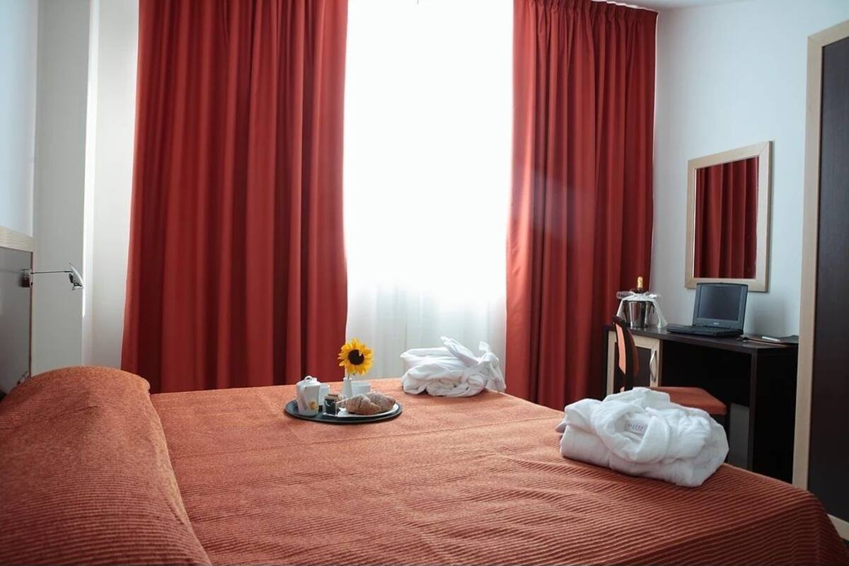 A hotel room with a bed, a desk with a laptop, a tray with breakfast items, and a window with red curtains.