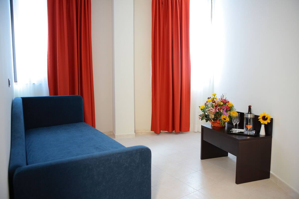 A room with a blue sofa, a dark wooden side table with a vase of flowers, and red curtains.