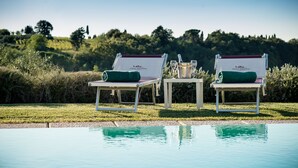 Deux fauteuils de jardin avec des coussins verts et une table sur laquelle se trouvent une bouteille et un verre, près d’une piscine offrant une vue dégagée sur un paysage vallonné.