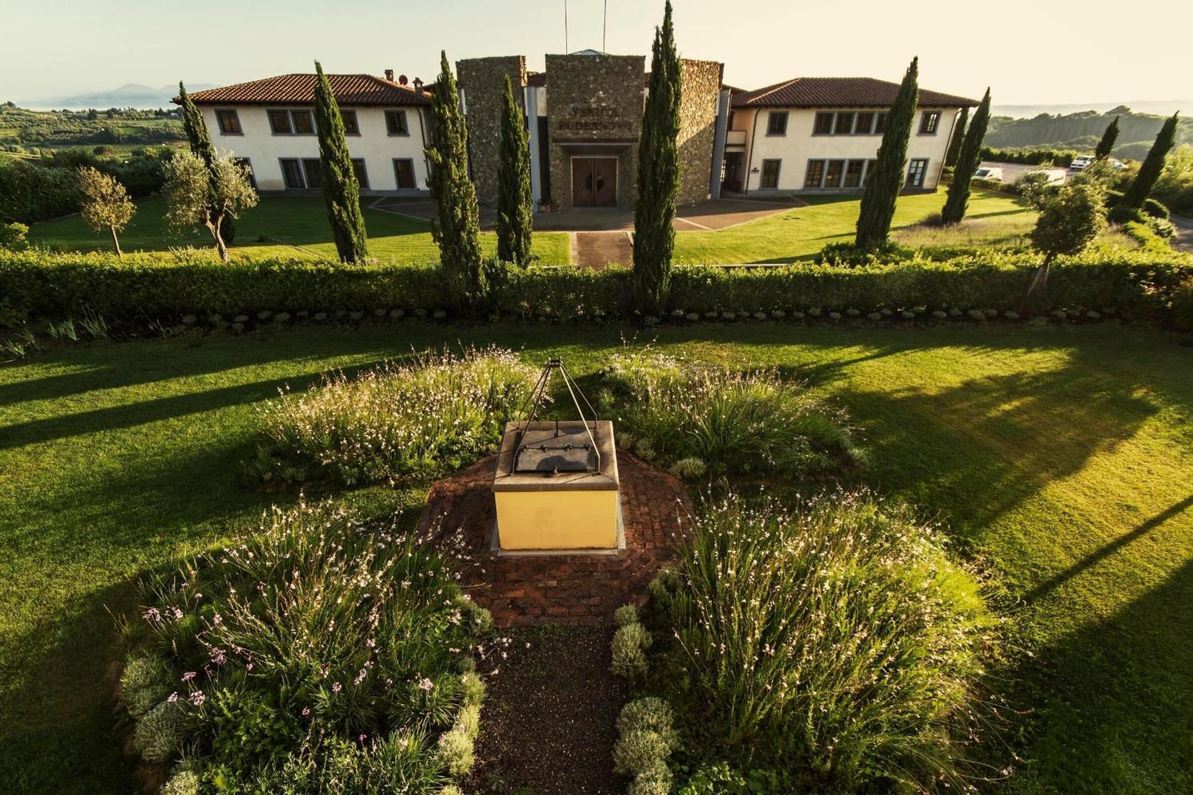Una casa grande con fachada de piedra, rodeada de jardines cuidados y altos cipreses.