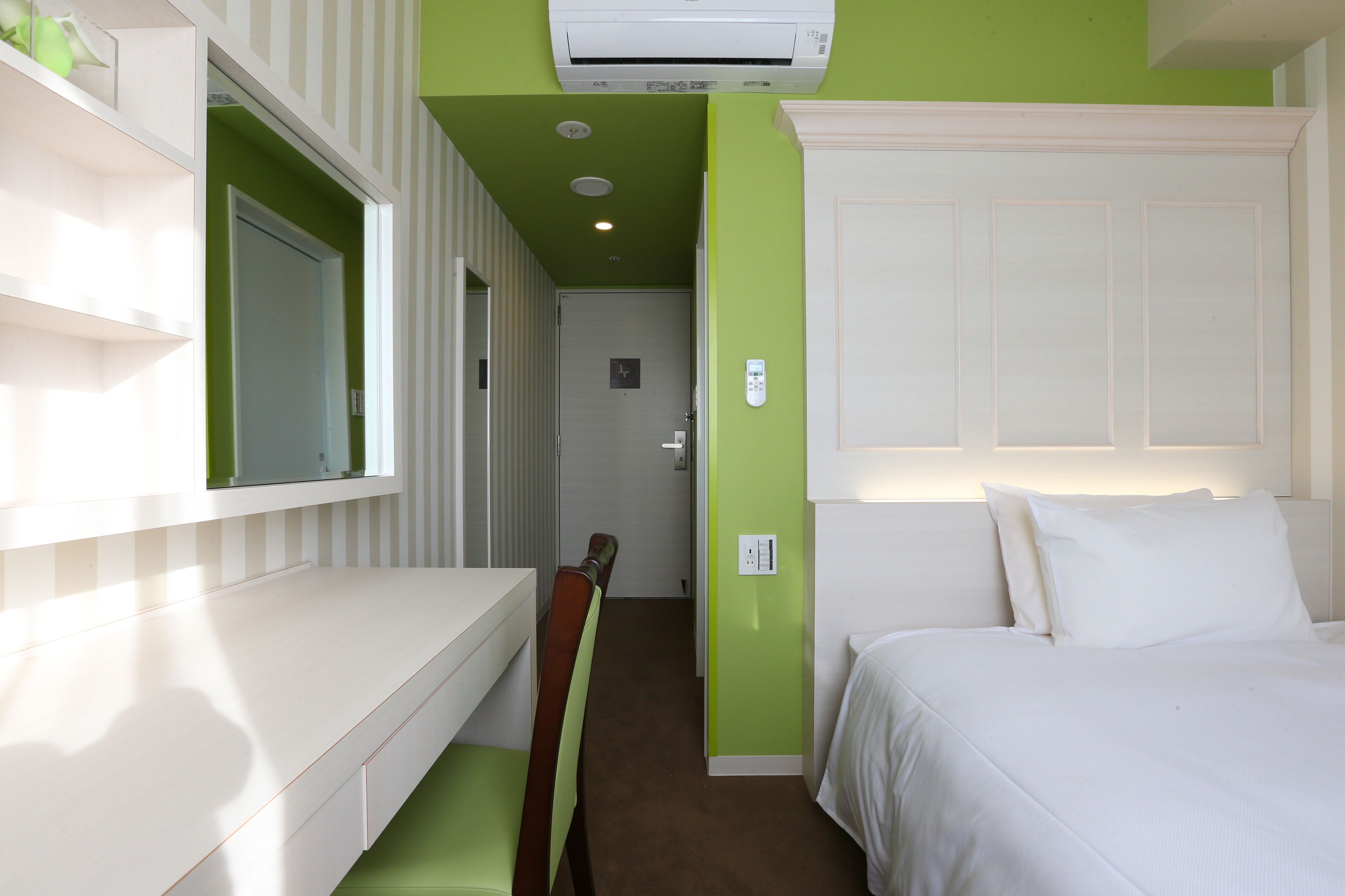 A hotel room with a white bed, green walls, a white desk, and a mirror.