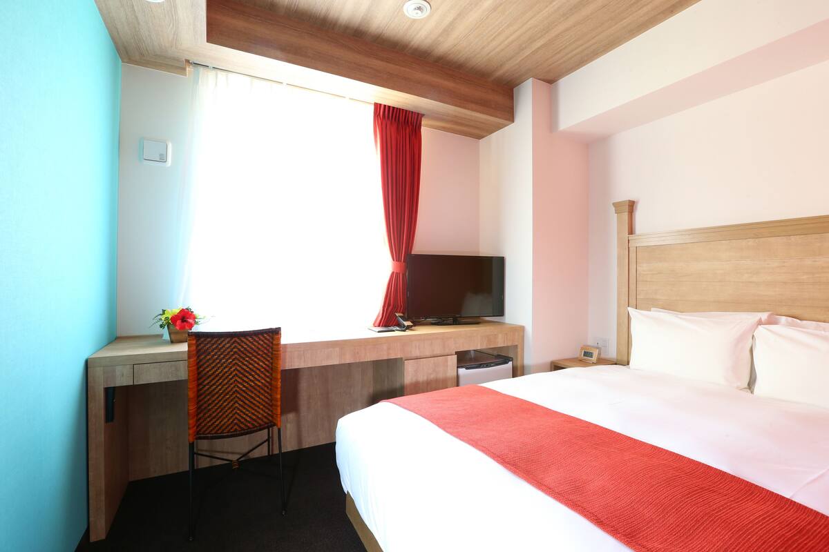 Ein Hotelzimmer mit Bett, Schreibtisch mit Computer, Stuhl, Fernseher und einem Fenster mit Vorhängen.