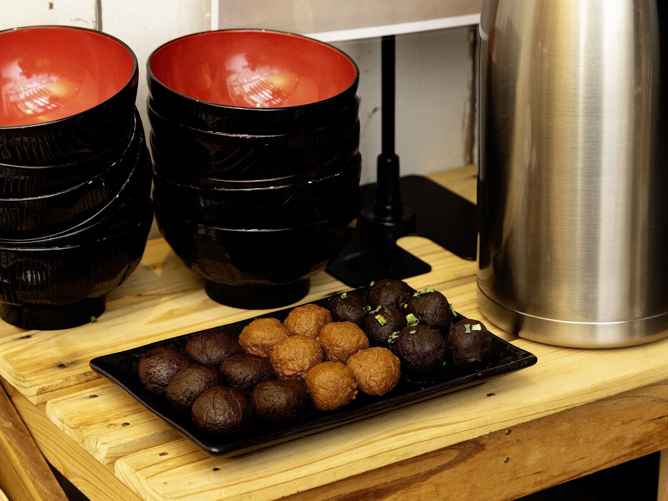 Une table en bois sur laquelle se trouve un plateau noir contenant des aliments de forme ronde et de couleur foncée, probablement des confiseries, ainsi qu’un récipient en acier inoxydable.