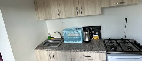 Une cuisine compacte avec des meubles de couleur bois clair, un plan de travail en granit, un évier en acier inoxydable, un égouttoir bleu, une machine à café et une cuisinière à gaz blanche.