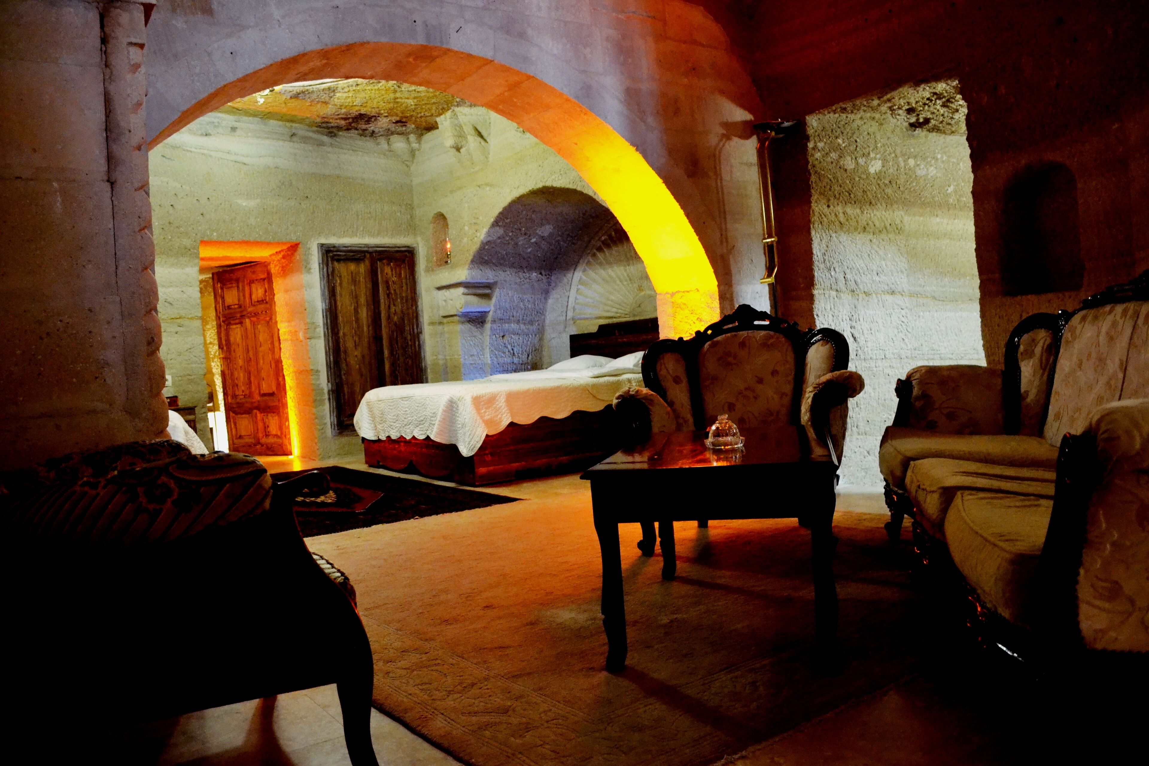 Una habitación tenuemente iluminada con una cama, dos sillones y una mesita. Un arco de entrada da a otra estancia.