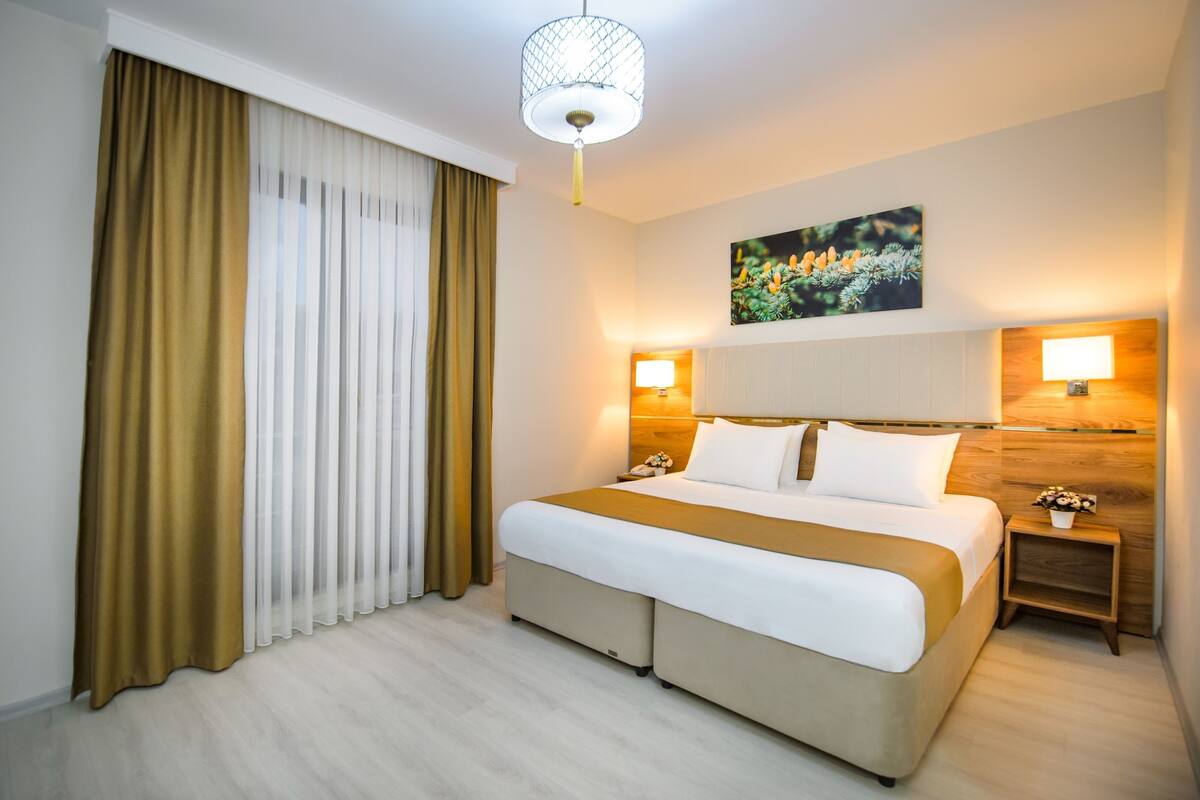 A hotel room with a large bed, two bedside tables, a wall-mounted lamp, and a window with curtains.