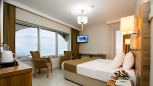 A hotel room with a bed, a desk, a chair, a TV, and a view of the sea. - White House Hotel (Trabzon)