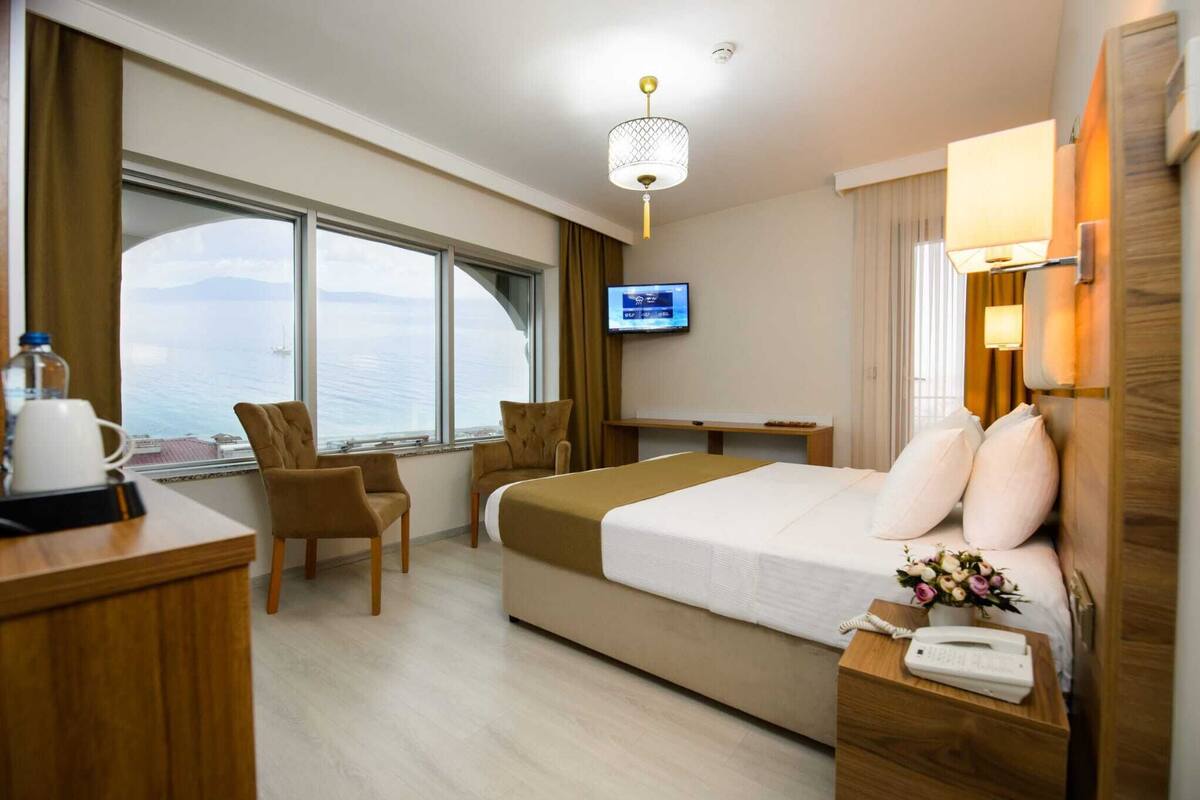 A hotel room with a bed, a desk, a chair, a TV, and a view of the sea.