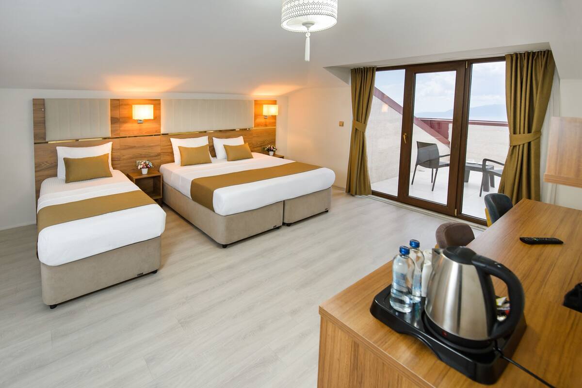 A hotel room with two beds, a desk with a kettle, and a view of the beach through large windows.