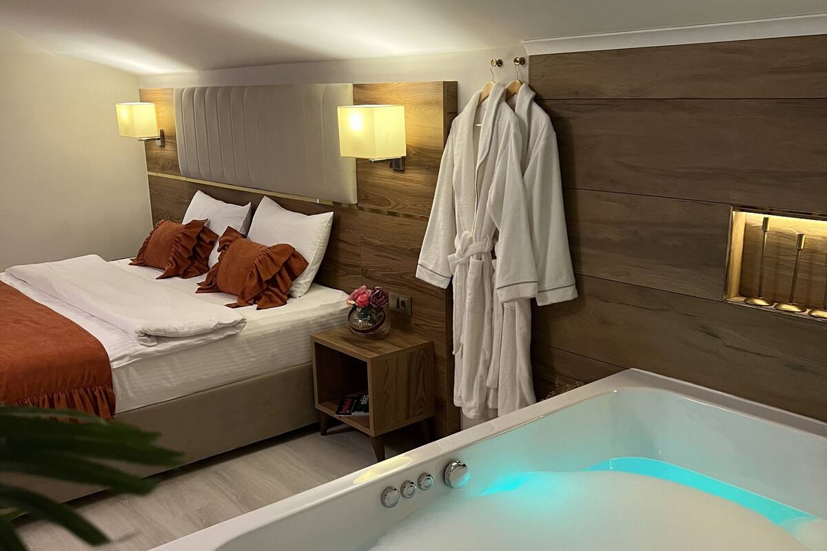 A modern hotel room with a large bathtub, a neatly made bed with brown pillows, a wooden headboard, and a small wooden nightstand with a lamp.