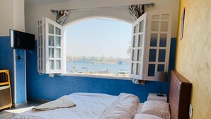 Quarto com cama, janela com persianas abertas, televisão, telefone e uma pequena mesa com abajur.