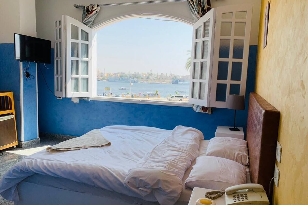 Ein Zimmer mit einem Bett, einem Fenster mit geöffneten Läden, einem Fernseher, einem Telefon und einem kleinen Tisch mit einer Lampe.