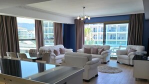 En moderne stue med en central ø, hvide sofaer og et stort vindue med gardiner.