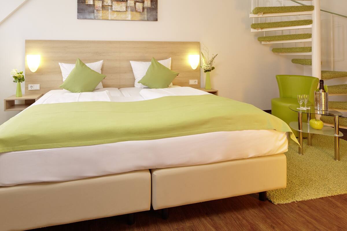A hotel room with a bed, green curtains, a green chair, a small table with a bottle and glasses, and a staircase with green steps.