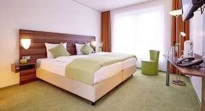 A hotel room with a large bed, bedside tables, a desk with a chair, a TV, and a window with curtains.