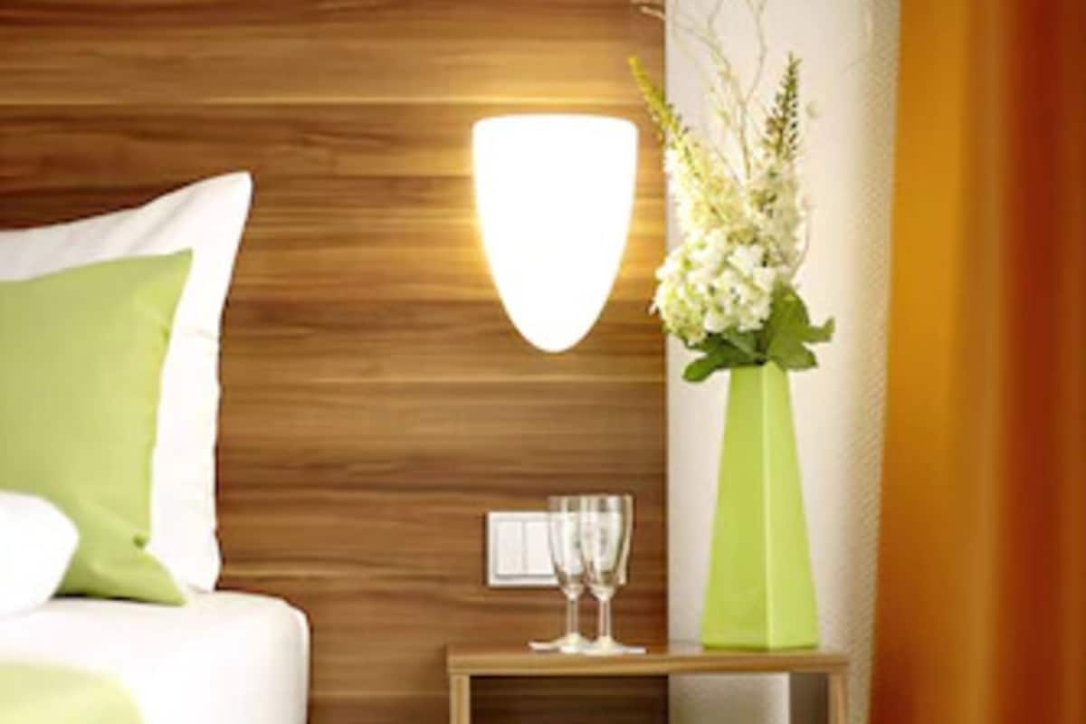 A modern hotel room with a wooden headboard, a green vase with flowers, a bedside table with a glass of water, and a green trash bin.