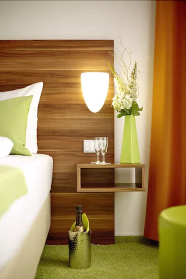 A modern hotel room with a wooden headboard, a green vase with flowers, a bedside table with a glass of water, and a green trash bin.