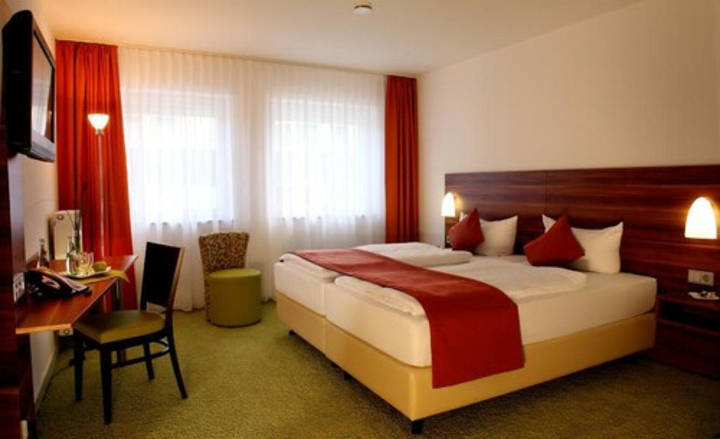 Ein Hotelzimmer mit einem großen Bett, einem Schreibtisch mit Stuhl, einem kleinen runden Hocker und einem Fernseher.