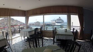 A restaurant with tables set for dining, a view of a cruise ship, and a cityscape in the background. - Surtel Hotel (Kusadasi)