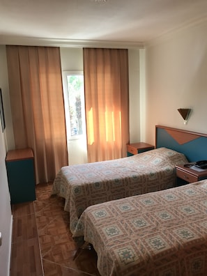 A hotel room with two beds, a wooden headboard, a nightstand, a small table, and a window with curtains.