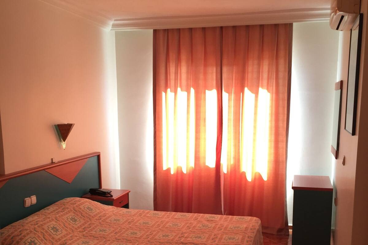 A bedroom with a bed, a nightstand, a wall-mounted lamp, and a window with curtains.