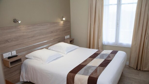 Una cama bien hecha con colcha blanca y marrón, cabecero de madera, mesitas de noche con lámparas y una ventana con cortinas.