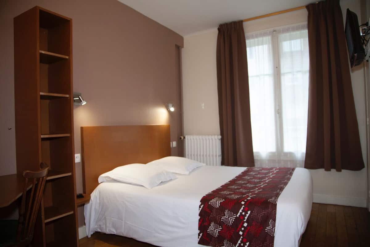 A hotel room with a bed, a wooden wardrobe, a desk, and a window with curtains.