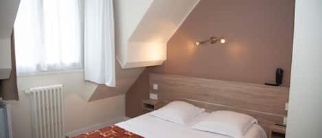 A bedroom with a sloped ceiling, a bed with white linens, a patterned throw blanket, a radiator, a window with sheer curtains, and a wall-mounted light fixture.